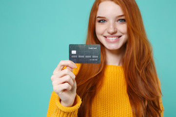 Cropped image of smiling young redhead woman girl in yellow sweater posing isolated on blue turquoise wall background in studio. People lifestyle concept. Mock up copy space. Hold credit bank card.