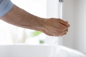 Side view close up young man put hands under running from tap water, cleaning skin in bathroom, morning routine. Responsible guy preventing spread virus infection, sanitizing hands regularly.