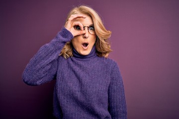 Middle age beautiful blonde woman wearing casual purple turtleneck sweater and glasses doing ok gesture shocked with surprised face, eye looking through fingers. Unbelieving expression.