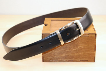 A stylish belt made of smooth, black leather and steel fittings lies beautifully on a wooden box, on a light background. Expensive gift option for men, women, premium leather, chic surprise