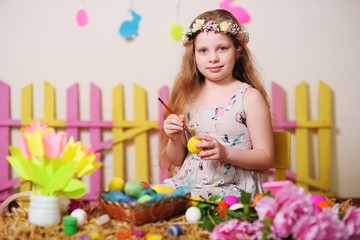 a child-a little girl in a smart dress and a wreath of flowers on her head smiles and paints Easter eggs on the background of the Easter decor