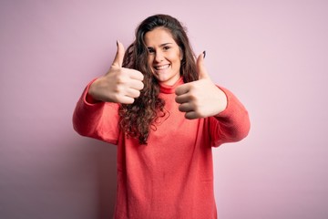 Young beautiful woman with curly hair wearing turtleneck sweater over pink background approving doing positive gesture with hand, thumbs up smiling and happy for success. Winner gesture.