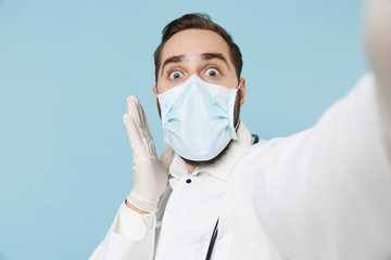 Close up of shocked male doctor man in medical gown face mask gloves isolated on blue background. Epidemic pandemic coronavirus 2019-ncov sars covid-19 flu virus concept. Doing selfie on mobile phone.