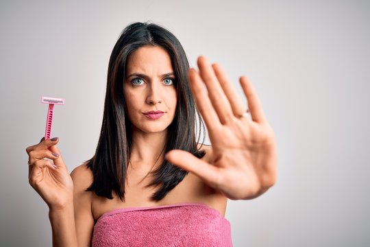Young brunette woman with blue eyes wearing bath towel using razor over white background with open hand doing stop sign with serious and confident expression, defense gesture