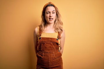 Young beautiful blonde woman wearing overalls and diadem standing over yellow background winking looking at the camera with sexy expression, cheerful and happy face.