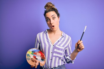 Fototapeta premium Young beautiful blonde artist woman painting using brush and palette over purple background scared in shock with a surprise face, afraid and excited with fear expression