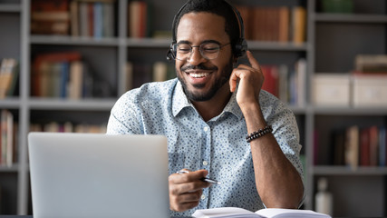 Smiling African American man in glasses and headset watch webinar on laptop making notes, happy...