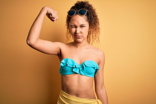 Young beautiful american woman on vacation wearing bikini over isolated yellow background Strong person showing arm muscle, confident and proud of power