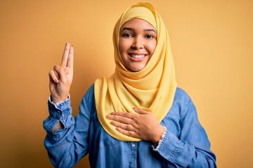 Young beautiful woman with curly hair wearing arab traditional hijab over yellow background smiling...
