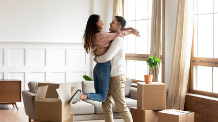 Young man lifting smiling mixed race wife, feeling excited about moving in new house. Happy family couple homeowners celebrating purchasing real estate, having fun in living room, relocation concept.