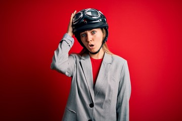 Young beautiful blonde motorcyclist woman wearing motorcycle helmet over red background surprised with hand on head for mistake, remember error. Forgot, bad memory concept.