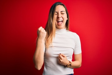 Fototapeta premium Beautiful blonde woman with blue eyes wearing casual white t-shirt over red background celebrating surprised and amazed for success with arms raised and eyes closed. Winner concept.