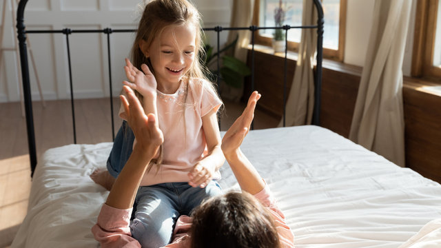 Happy little girl sitting on mommy's belly, playing patty cake in bedroom. Playful small adopted child clapping hands with joyful foster parent, having fun enjoy family weekend together on bed.