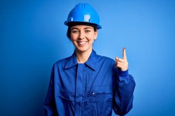 Young beautiful worker woman with blue eyes wearing security helmet and uniform pointing finger up with successful idea. Exited and happy. Number one.