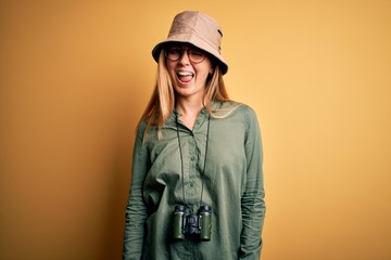 Beautiful blonde explorer woman with blue eyes wearing hat and glasses using binoculars winking...