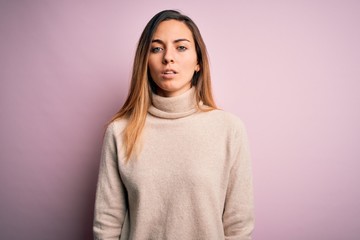 Beautiful blonde woman with blue eyes wearing turtleneck sweater over pink background with serious expression on face. Simple and natural looking at the camera.