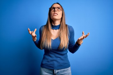Young beautiful blonde woman with blue eyes wearing glasses standing over blue background crazy and mad shouting and yelling with aggressive expression and arms raised. Frustration concept.