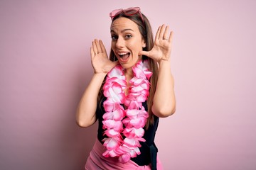Young beautiful blonde woman wearing swimsuit and floral Hawaiian lei over pink background Smiling cheerful playing peek a boo with hands showing face. Surprised and exited