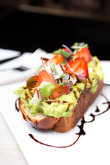 A stylized slice of avocado toast with tomatoes and strawberries, in a restaurant or kitchen setting.