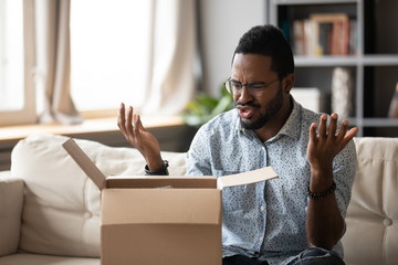 Unhappy African American man client disappointed with product quality shopping online, mad biracial male open cardboard package frustrated with wrong Internet order, bad delivery service concept - obrazy, fototapety, plakaty