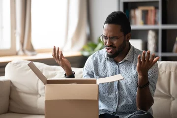 Fotobehang Unhappy African American man client disappointed with product quality shopping online, mad biracial male open cardboard package frustrated with wrong Internet order, bad delivery service concept © fizkes