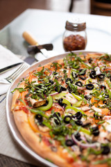 A rustic pizza pie with meat, veggies and basil toppings, in a restaurant or kitchen setting.