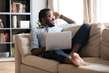 Smiling African American millennial man relax on comfortable couch in living room work on laptop...