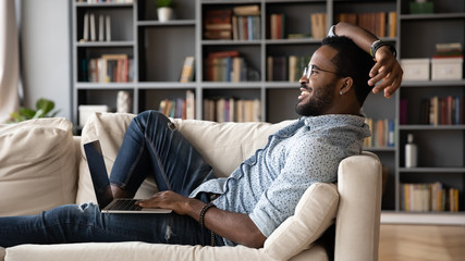 Happy biracial young man relax on comfortable sofa at home working on modern computer gadget,...