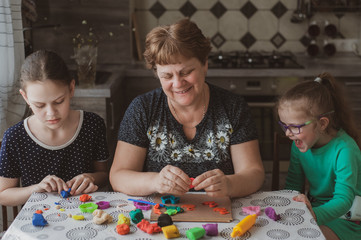An adult woman and two little girls make plasticine figurines at home. Grandmother and granddaughters are sitting at home. Family Values Concept