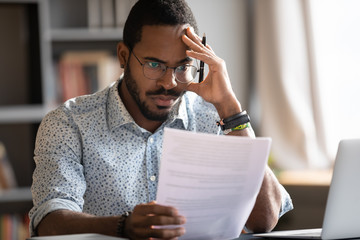 Pensive unhappy biracial man in glasses feel distressed reading bad news in paperwork letter,...