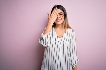 Young beautiful woman wearing casual striped t-shirt and glasses over pink background covering one eye with hand, confident smile on face and surprise emotion.