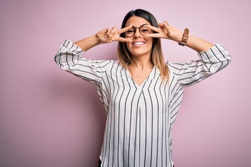 Young beautiful woman wearing casual striped t-shirt and glasses over pink background Doing peace symbol with fingers over face, smiling cheerful showing victory