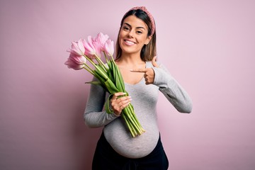 Young beautiful brunette woman pregnant expecting baby holding bouquet of tulips very happy pointing with hand and finger