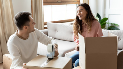 Happy mixed race woman talking with smiling husband while packing home stuff into carton boxes for moving in new house. Joyful family couple preparing belongings for delivery, relocation service.