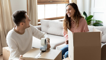 Happy mixed race woman talking with smiling husband while packing home stuff into carton boxes for...