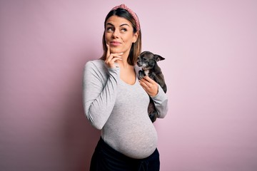 Young beautiful brunette woman pregnant expecting baby holding chihuahua puppy serious face thinking about question, very confused idea