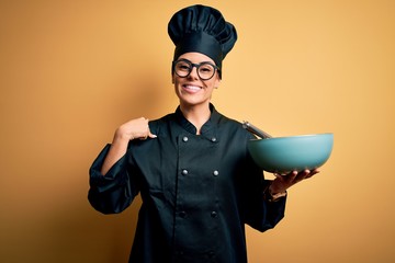 Young beautiful brunette chef woman wearing cooker uniform and hat holding bowl and whisk with surprise face pointing finger to himself