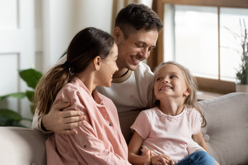 Happy young man cuddling sitting on couch smiling mixed race wife and little preschool daughter. Excited joyful woman and small child girl looking at father husband, listening to good news at home.