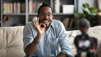 Millennial african American male blogger or tutor sit on couch shooting video blog on modern web cam, young biracial man coach in glasses talk with subscribers record tutorial or training course - obrazy, fototapety, plakaty