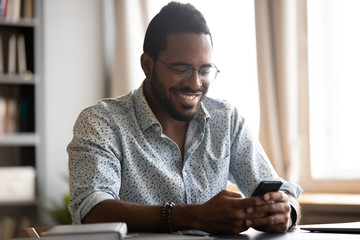 Happy biracial man in glasses sit at desk have fun texting messaging using wireless internet on smartphone gadget, smiling African American male in spectacles laugh watch funny video on cellphone - obrazy, fototapety, plakaty