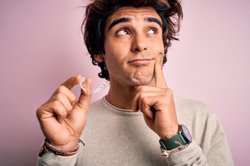 Young handsome man holding aligner standing over isolated pink background serious face thinking about question, very confused idea