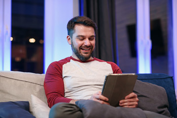 Slow motion of likable modern good-natured 30-aged man with well-groomed beard which resting on the sofa at home and browsing apps on tablet pc with lucky smile