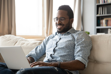 Smiling African American millennial man freelancer sit on couch in living room working on modern...