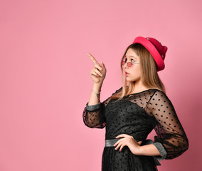 Teenage girl in sunglasses, black dress and red hat. She pointing at something by forefinger, posing on pink background. Close up