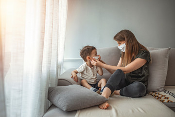 Obraz na płótnie Canvas Side view of adult woman putting on medical mask on little boy during coronavirus outbreak against gray background. Mother and her son putting masks on protect themselves from viruses