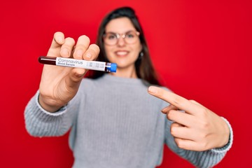 Fototapeta premium Young beautiful girl wearing glasses holding laboratory blood sample of coronavirus disease very happy pointing with hand and finger