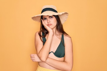 Young beautiful girl wearing swimwear bikini and summer sun hat over yellow background thinking looking tired and bored with depression problems with crossed arms.
