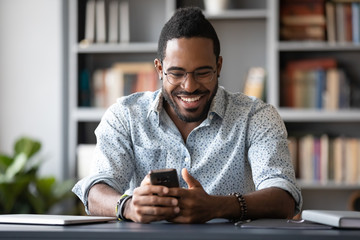 Overjoyed millennial African American man in glasses sit at desk laugh texting or typing on cellphone, happy biracial male in spectacles have fun watch funny video on smartphone gadget indoors - obrazy, fototapety, plakaty