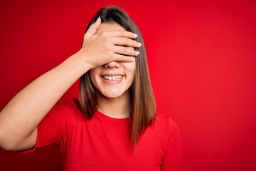 Young beautiful brunette girl wearing casual t-shirt over isolated red background smiling and laughing with hand on face covering eyes for surprise. Blind concept.