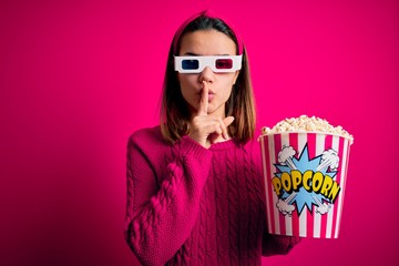 Obraz premium Young beautiful girl watching movie using 3d glasses eating box with popcorns asking to be quiet with finger on lips. Silence and secret concept.